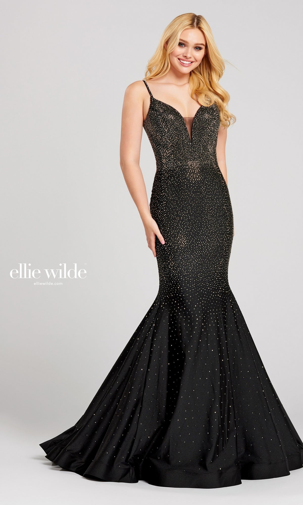 A woman wears the Ellie Wilde Beaded Mermaid Prom Dress with Cut Out—fitted, black, floor-length, with thin straps and sparkling embellishments—standing and smiling against a plain background.